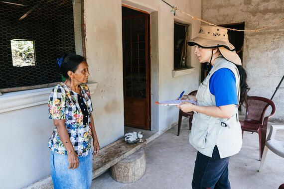 A woman in an OCHA vest talks to a woman facing her. They are standing in a verandah of a house.