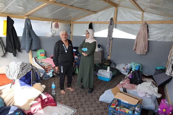 A woman and a man stand inside a tent with packages and clothes and other household goods.