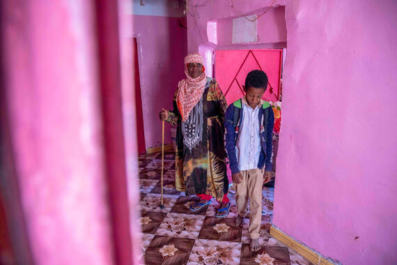 A woman and a young boy stand inside a house near a doorway. The walls of the house are a bright pink.
