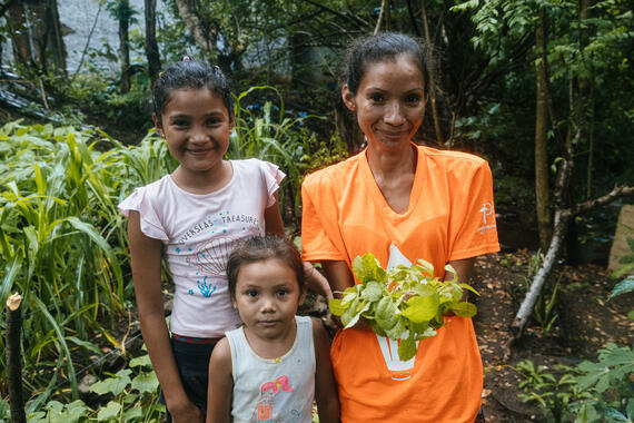 A woman stands in a garden holding some leaves in her hands. Two young girls stand next to her smiling.