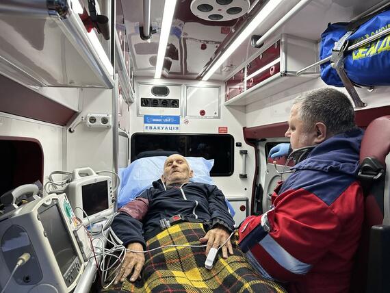 A man lies on a bed inside an ambulance hooked up to monitors. A person a in red vest is seated beside him.