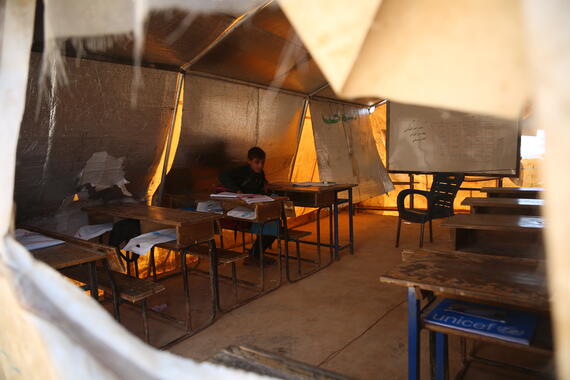 Aboy sits behind a desk in a tent. There are several benches and desks inside the tent.