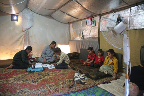 A man seated on a mat inside a tent looks on as two boys seated near him focus on notebooks on the mat. Other children are seated near them.