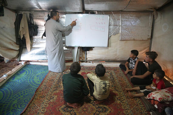 A standing man writes on a blackboard inside a tent. Children are seated on a mat below.