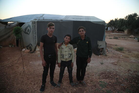Three boys stand outside in front of a tent.