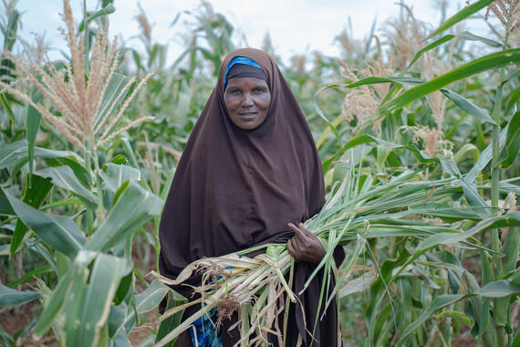 A smiling woman stands midst a maize field holding a flowering maize plant.