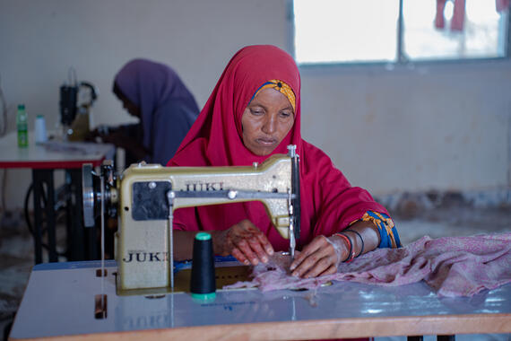 A woman sits behind a sewing machine, stitching a garment.