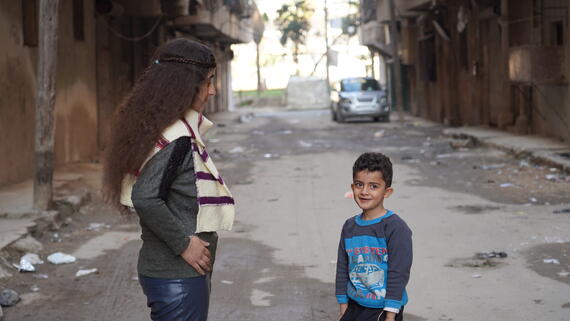 A young woman faces a boy, who is facing the camera smiling. They stand on a quiet road lined by buildings.