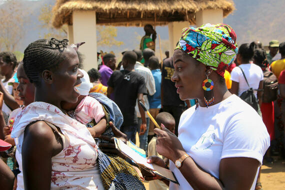 A woman talks to another in a n open space outside. Crowds of people can be seen in the background.
