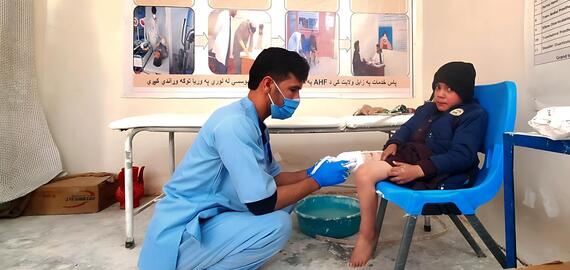 A man in blue clothes examines the bandaged leg of a young boy seated on a chair in Afghanistan.