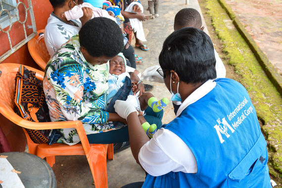 A woman wearing a blue vest that has International Medical Corps printed on it vaccinates a baby in the arms of a seated woman.