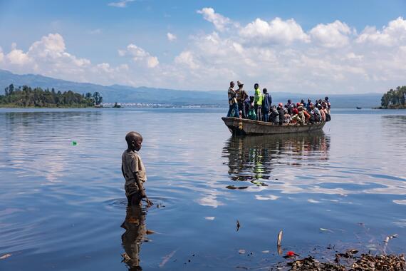 A young boy stands by the water looking at a small, overcrowded boat approaching the shore in DRC