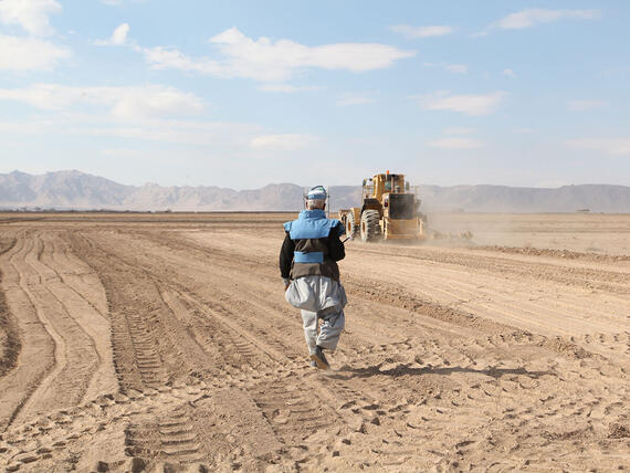 A man with a mask is walking across an open field. A machine can be seen in the background.