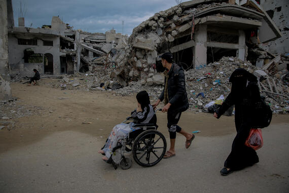 A young boy pushes a young girl ina wheel chair on an untarred road. Debris and destroyed buildings can be seen in the background.