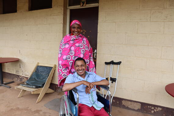 A young man sits on a wheelchair smiling and a smiling woman stand behind teh wheelchair. Crutches placed against a wall of a building can be seen behind them.
