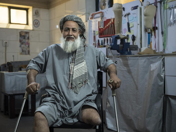 An older man sits on a chair with crutches. One of his legs is missing.